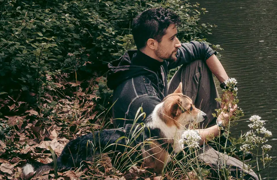 Camille Proust, assis au bord de l’eau avec sa chienne Plùm dans un environnement végétalisé.